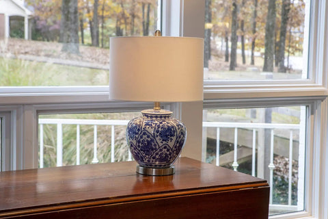 Blue And White Floral Ceramic Table Lamp With White Linen Shade And Brushed Nickel Accents 3 Way Lighting Ginger Jar Inspired Cozy Decor For Living Room Or Bedroom Soft Warm Glow And Timeless Charm on a wooden table by window.