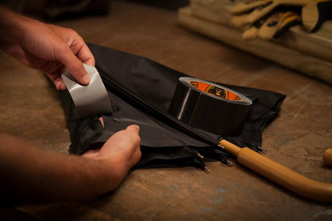 Hands using Heavy Duty Double Thick Black Duct Tape 1.88 Inch By 12 Yard Roll on black fabric, showing repair use.