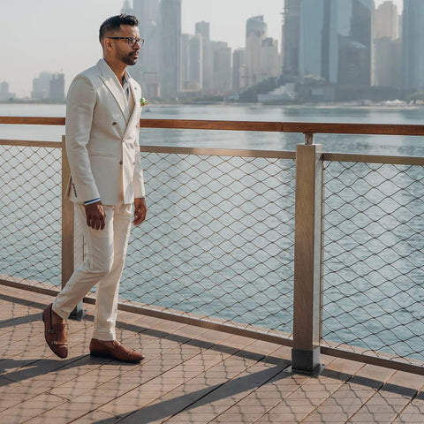 Man in a beige suit walking on a deck by the waterfront city skyline.