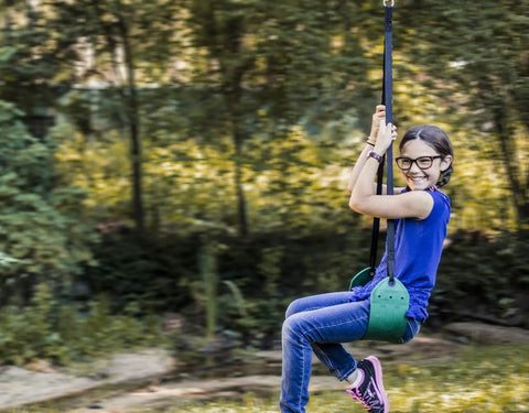 Child riding Green Zip Line Swing Seat With Nylon And Powder Coated Alloy Steel Build 250 Pound Weight Limit 40 Inch Height outdoors