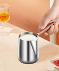 Hand holding Turkish Coffee Pot Stainless Steel Multipurpose Milk Frothing Pitcher And Coffee Kettle for Latte Art on table.