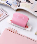 Six-Hole Binding Machine For Planners, White Manual Punch For A4 B5 A5 Paper, pink handle, on a desk with notebooks