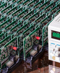Rows of green electronic circuit boards with red components and a digital testing device next to them.