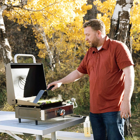 Man using Stainless Steel Propane Gas Portable Flat Top Griddle Station For Outdoor Kitchens Camping Tailgate Ready And Tabletop Cooking 17 Inch 12,000 BTU Heavy Duty Easy Cleanup Versatile Breakfast And Burgers on picnic table