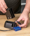 Person connecting battery to Airshield Pro Remote Battery Charging Cradle For AIR/P/7 on wooden table, showing charging process.