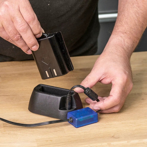 Person connecting battery to Airshield Pro Remote Battery Charging Cradle For AIR/P/7 on wooden table, showing charging process.