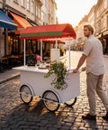 Hustle Made Easy With This Heavy Duty Outdoor Vending Cart, Weatherproof Canopy Design, Smooth Ride Wheels, Huge Prep Counter, And Eye Catching Stage Setup For Your Small Business Side Hustle, being pushed on a cobblestone street.