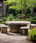 Outdoor patio with stone table and wooden stools in garden setting with lush greenery and cobblestone flooring.