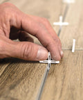 Hand placing 1/8 Inch Hard Tile Spacers for Traditional or Leave-In Installations, 300 Pack, ensuring consistent grout joints on wood-look tiles.
