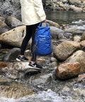 Person crossing rocky stream holding fully waterproof hiking backpack with 25L capacity, lightweight packable travel daypack in teal.