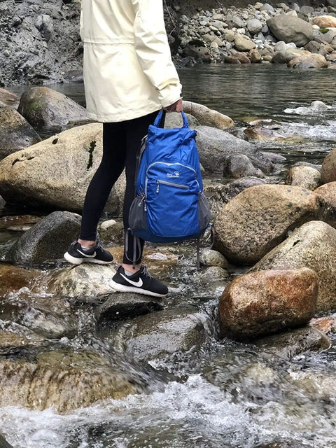 Person crossing rocky stream holding fully waterproof hiking backpack with 25L capacity, lightweight packable travel daypack in teal.