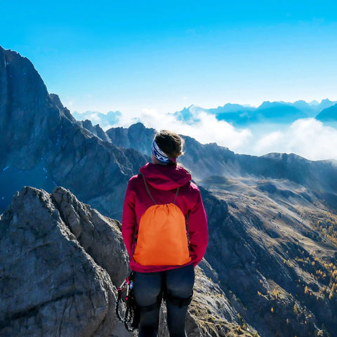 Person hiking with an orange 10 Color Drawstring Backpack For Gym And Travel, Lightweight Yet Durable Polyester Sack Pack, on mountain trail.