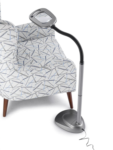 MultiFlex LED Floor Magnifier Lamp With Ultra Wide 6 Inch Viewing Area next to chair, silver finish, adjustable neck