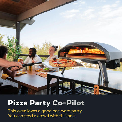 Koda 16 Natural Gas Pizza Oven for Outdoor Cooking, showing a pizza baking on the black durable steel body, with people dining.