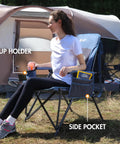 Woman sitting on oversized folding high back lawn chair for adults with cup holder, carrying strap, hard armrests, 400Lb capacity, blue.