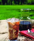 Glass of iced coffee with swirling cream on a table with coffee beans and a carafe, outdoors in a park setting.