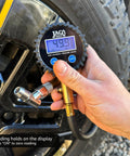 Close-up of JACO ElitePro digital tire pressure gauge at tire valve; blue LCD displays 49.9 PSI.