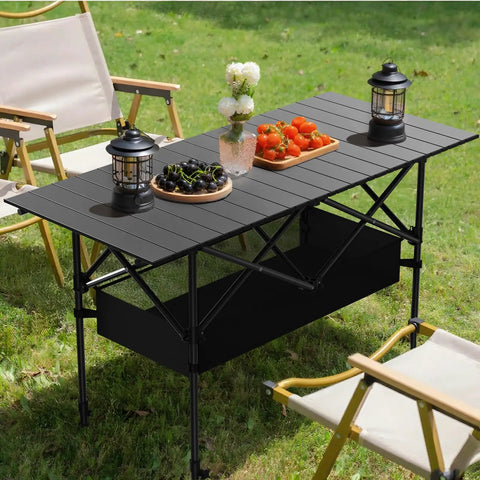 4Ft Folding Camping Table With Carry Bag And Mesh Storage in black, set on grass with lanterns, fruits, and flowers.