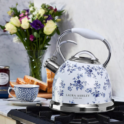 Floral design stovetop kettle with whistle, white and blue pattern, on gas hob, next to tea set and flowers in kitchen.