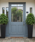Front view of Resin Tall Planters For Outdoor Plants Set Of 2 Large Tapered Flower Pots With 4 Drainage Holes No Fuss Modern Outdoor Decor For Porch Patio Backyard Home Garden Black 24 Inch placed by a door.