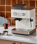 Semi Automatic Espresso Machine With Built In Burr Grinder And Smart Dosing Tech For Smooth One Or Two Shot Pulls Sturdy Metal Body In Porcelain White being used to tamp coffee.