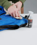 Person applying Outdoor Gear UV Protectant and Conditioner Spray for Plastic, Vinyl, and Neoprene on blue fabric.