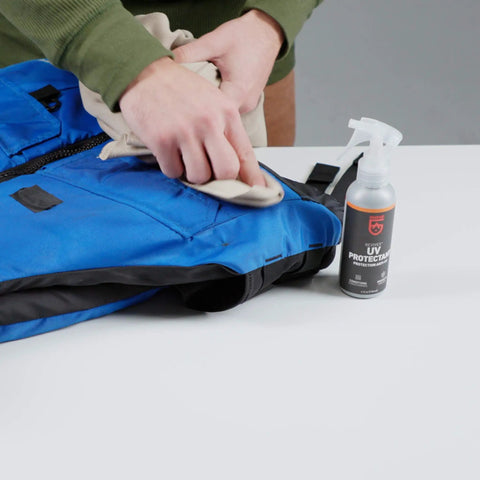 Person applying Outdoor Gear UV Protectant and Conditioner Spray for Plastic, Vinyl, and Neoprene on blue fabric.
