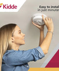 Woman installing hardwired smoke and carbon monoxide detector with battery backup and interconnectable design for whole home safety.