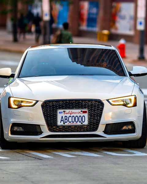 Front view of a white car on the street with a prominently attached Front License Plate Bracket With Full Border In Black ABS.