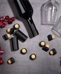 100 Pack Black Wine Bottle Sealers With Solid Airtight Plastic Cork Tops scattered around a wine bottle and grapes.