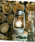 Oil lamp with glowing wick, silver metal frame, standing on cut logs. 10 Piece Cotton Wick Set For Oil Lamps Kerosene And Propane Lanterns