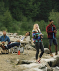 Group of friends fishing by a lake with trees in the background, some sitting near a campfire, others standing with fishing rods.