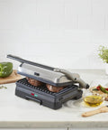 Cuisinart Griddler Indoor Grill And Panini Press With Removable Nonstick Plates cooking burgers on gray grill, with veggies and oil nearby.