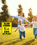 3 Pack Slow Down Kids At Play Signs 14.2 By 11 Inches Double Sided Weatherproof Yard Street Safety Sign Yellow High Visibility For Neighborhoods And Outdoor Use shown in a park setting with children playing