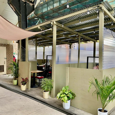 Outdoor seating area with corrugated roof, potted plants, and semi-private dining booths on a sidewalk.