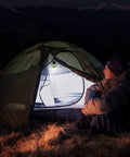 Person sitting by a tent lit with Rechargeable Camping Light With Clip Hook Your Go To Portable Tent Lantern 3 Lighting Modes High Low Strobe Hanging LED Bulb For Hiking Fishing Backpacking Emergency Power Outages And Outdoor Adventures in the mountains at night.