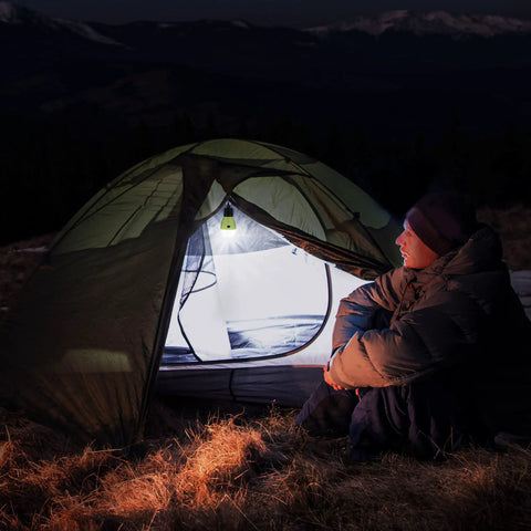 Person sitting by a tent lit with Rechargeable Camping Light With Clip Hook Your Go To Portable Tent Lantern 3 Lighting Modes High Low Strobe Hanging LED Bulb For Hiking Fishing Backpacking Emergency Power Outages And Outdoor Adventures in the mountains at night.