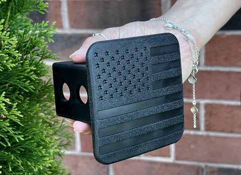 Hand holding Patriotic 2 Inch Tow Hitch Cover Plug With American Flag Design, Heavy Duty Trailer Receiver Cap, textured black surface.