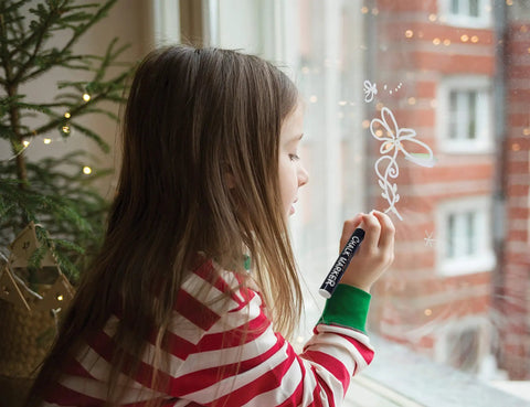 Child drawing on a window with chalk marker, focused on creativity, indoors, wearing striped pajamas, festive atmosphere visible.