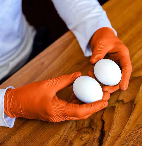 Hands wearing Orange Lightning Gloves OR-L Large holding eggs on a wooden surface, suitable for food service.