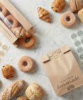Clear Window Bread Bags For Homemade Bread Keep Sourdough Fresh For Days And Make Gifting Your Bakes A Breeze Eco Friendly Kraft Paper Packaging For Bakery Goods shown with various breads and stickers on marble surface.