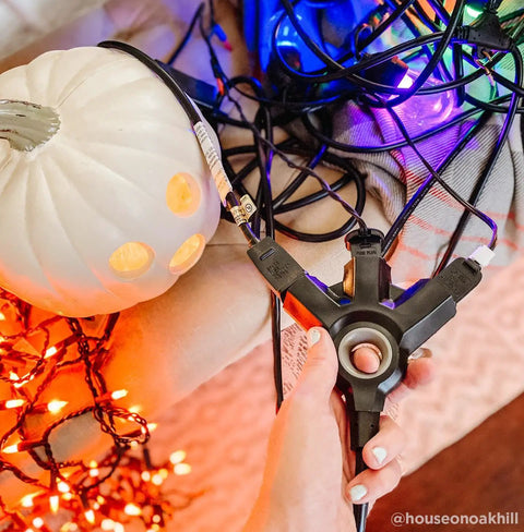 Hand holding EZGrip 25 Ft Heavy Duty Outdoor Extension Cord With Multiple Outlets among Halloween lights and a white pumpkin.
