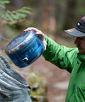 Person placing Adjustable Bear Canister For Backpacking And Camping into a grey backpack in a forest setting.