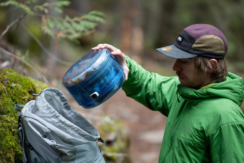 Person placing Adjustable Bear Canister For Backpacking And Camping into a grey backpack in a forest setting.