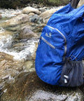 Fully Waterproof Hiking Backpack With 25L Capacity, Lightweight Packable Travel Daypack in teal by a stream, roll top closure visible.