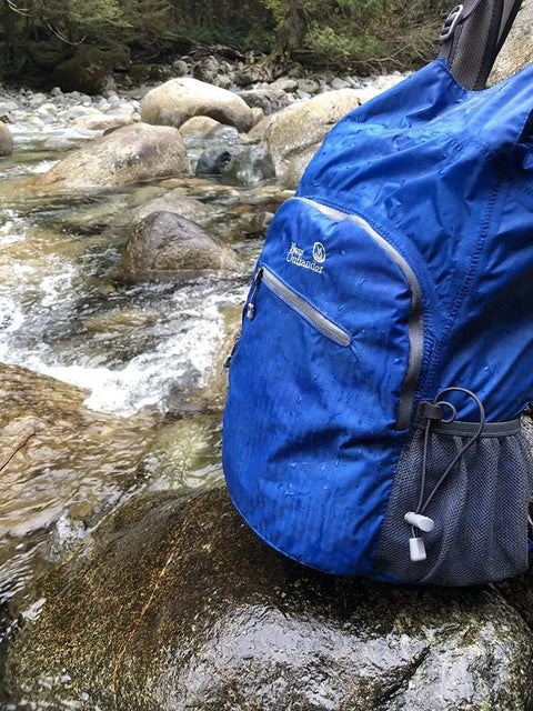 Fully Waterproof Hiking Backpack With 25L Capacity, Lightweight Packable Travel Daypack in teal by a stream, roll top closure visible.