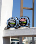 Pair of Komelon tape measures with high visibility labels, 33 ft and 25 ft sizes, set against urban background with blue sky.