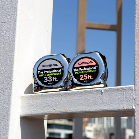 Pair of Komelon tape measures with high visibility labels, 33 ft and 25 ft sizes, set against urban background with blue sky.
