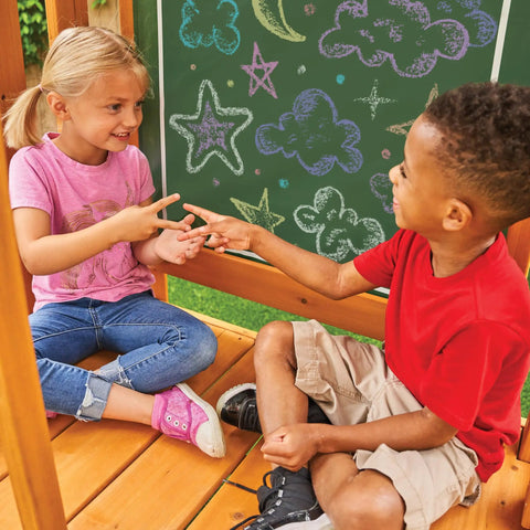 Children playing on a weatherproof wooden swing set for backyard with UV and mold protection, featuring a vinyl chalk wall.
