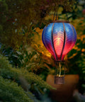 Blue Purple Hot Air Balloon Solar Lantern for Garden Decor lit with flickering flame hanging among greenery outdoors at night